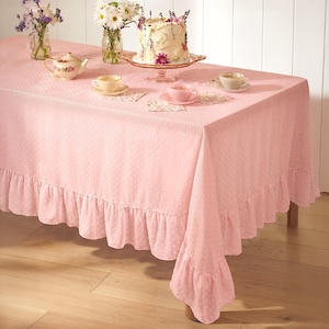 May include: A pink tablecloth with a ruffled edge and white polka dots covers a table set for tea. A cake on a pedestal, teacups, and a teapot are arranged on the table, with vases of flowers.