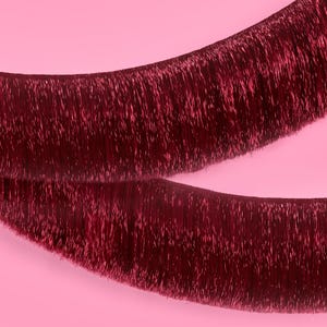 May include: Two strands of burgundy tinsel, slightly curved, against a soft pink background. The tinsel has a shimmering, metallic appearance, with individual strands visible. The image is a close-up, highlighting the texture and color of the decorative material.