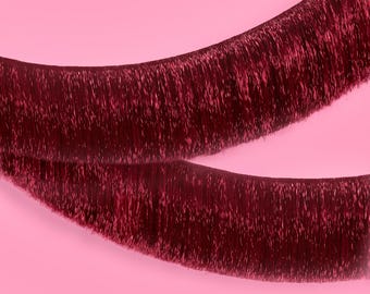 May include: Two strands of burgundy tinsel, slightly curved, against a soft pink background. The tinsel has a shimmering, metallic appearance, catching the light and creating a textured effect. The image is a close-up, highlighting the detail of the strands.