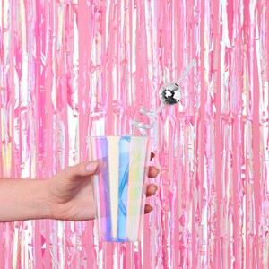 K&ouml;nnte beinhalten: Eine Hand h&auml;lt ein klares, irisierendes Plastikglas mit einer silbernen Discokugel an einem Strohhalm. Der Hintergrund ist ein rosa, irisierender Folienvorhang.