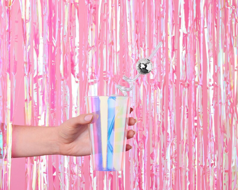 K&ouml;nnte beinhalten: Eine Hand h&auml;lt ein klares, irisierendes Plastikglas mit einer silbernen Discokugel an einem Strohhalm. Der Hintergrund ist ein rosa, irisierender Folienvorhang.