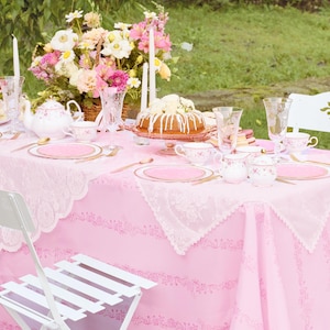 May include: A pink tablecloth is set for a tea party, complete with floral arrangements, a cake, and a tiered serving tray. White chairs with floral cutouts and delicate china with gold trim complete the scene. The table is set with pink plates and gold utensils.