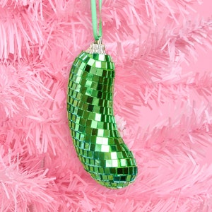 May include: A green, pickle-shaped Christmas ornament covered in small, reflective squares hangs from a green ribbon against a pink Christmas tree. The ornament has a silver cap and is a festive holiday decoration.