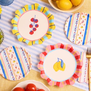 May include: A collection of colorful, scalloped-edge plates with fruit and floral designs. The plates feature illustrations of tomatoes, lemons, and striped patterns in shades of pink, yellow, and blue. The plates are arranged on a blue and white striped tablecloth.