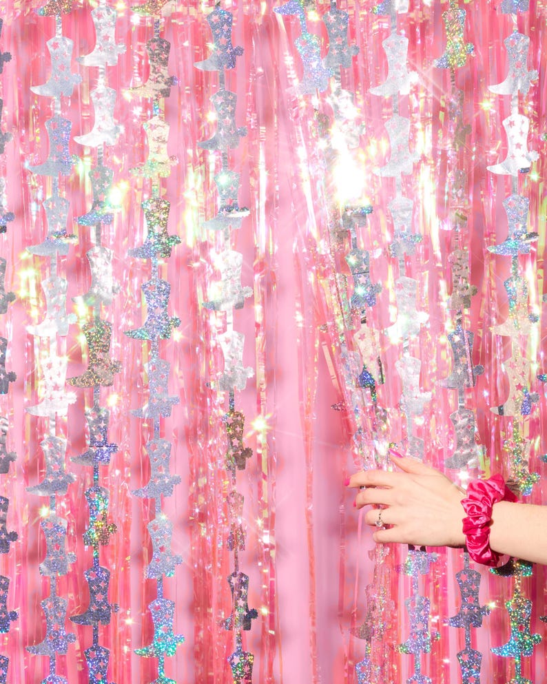 May include: Pink and silver iridescent foil fringe curtain with a boot shape cut out. The curtain is hanging from a wall and a hand is reaching through the curtain.