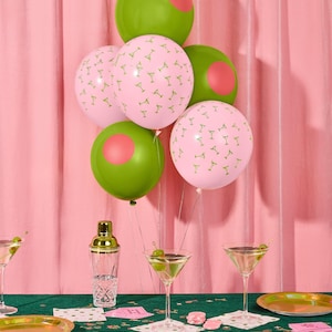 May include: A festive party scene with green and pink balloons. Some balloons are pink with martini glass patterns, others are green with pink circles. Martini glasses, gold plates, and a cocktail shaker are on a green tablecloth.