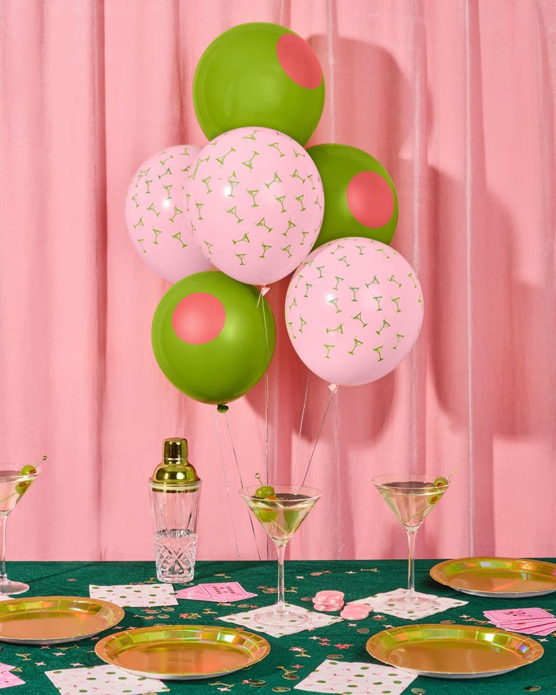 May include: A festive party scene with green and pink balloons. Some balloons are pink with martini glass patterns, others are green with pink circles. Martini glasses, gold plates, and a cocktail shaker are on a green tablecloth.