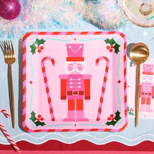 May include: A festive Christmas table setting with a pink square plate featuring a nutcracker design, candy canes, and holly. Gold cutlery and a matching napkin complete the holiday theme. A pink ornament and tinsel are in the background.