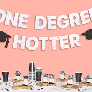 May include: A graduation party banner with iridescent letters that read "One Degree Hotter". The banner is decorated with black graduation caps. The banner is hanging above a table with cupcakes decorated with graduation caps and the words "One Degree Hotter" and "It's a graduation".