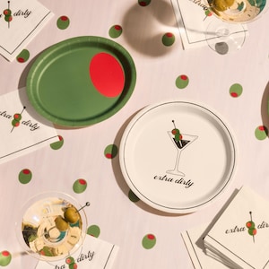 May include: A festive party scene with green and red olive-shaped confetti scattered around. White napkins and plates feature a martini glass illustration and the words "extra dirty." Green oval plates with red accents are also present.