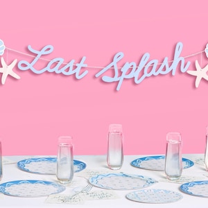 May include: A party table setting with a pink background. A banner that says "Last Splash" is strung across the top, decorated with seashells and starfish. The table is set with blue and white plates, glasses, and napkins.