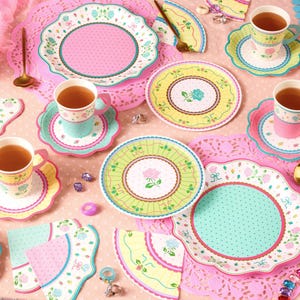 May include: A whimsical tea party setting with pastel-colored plates, cups, and saucers. The plates feature floral and polka dot designs in pink, green, and yellow. Gold spoons and forks are arranged around the table, with pink doilies and tea cups filled with tea.