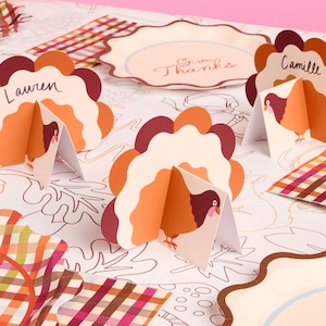 May include: Thanksgiving-themed place cards shaped like turkeys, with names written on them. The cards are in shades of orange, brown, and cream, with a turkey illustration. Decorative plates and patterned napkins are also visible.