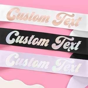 May include: Three satin sashes in white, black, and lavender with the text "Custom Text" in a glittery, cursive font.