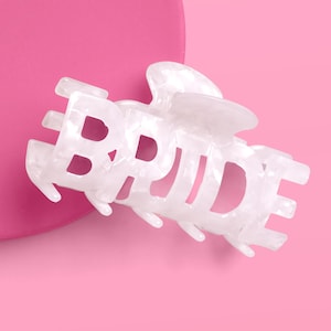 May include: White marbled-effect hair claw clip spelling out the word "BRIDE". The clip is set against a pink background. This bridal hair accessory is perfect for a wedding or bachelorette party.