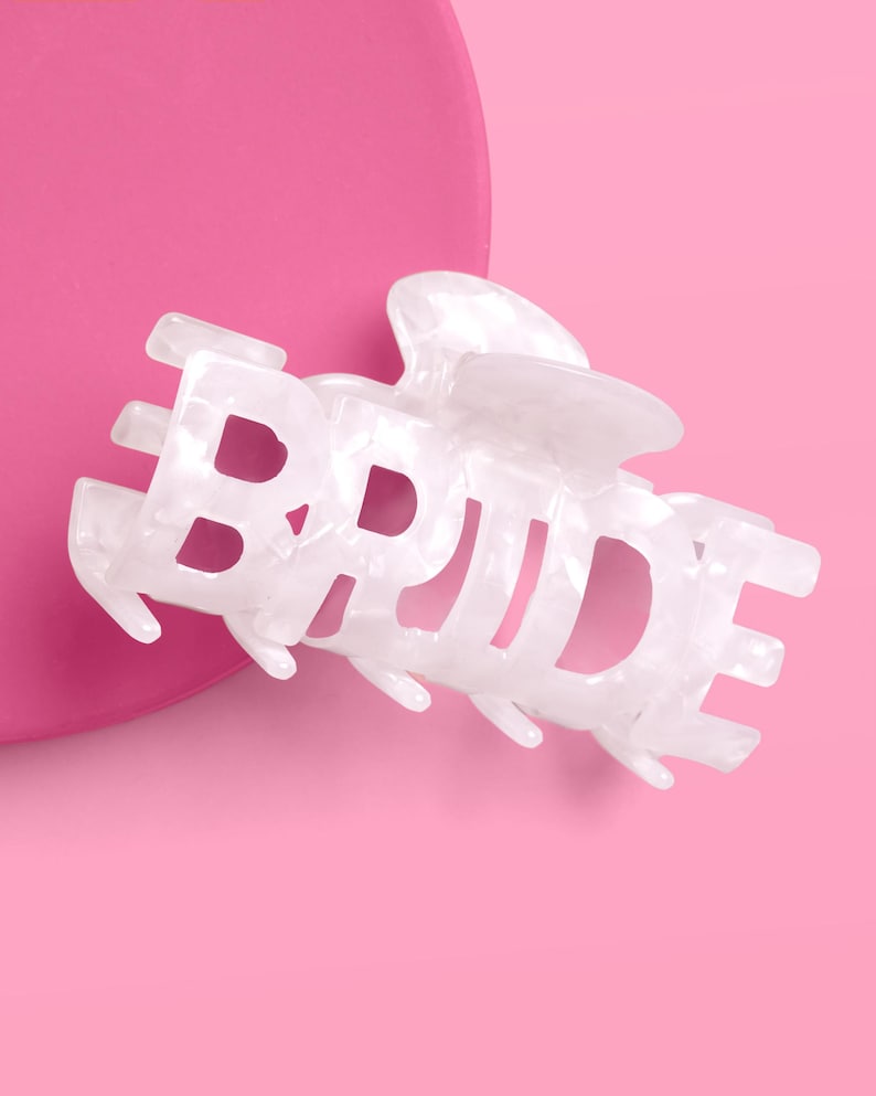 May include: White marbled-effect hair claw clip spelling out the word "BRIDE". The clip is set against a pink background. This bridal hair accessory is perfect for a wedding or bachelorette party.