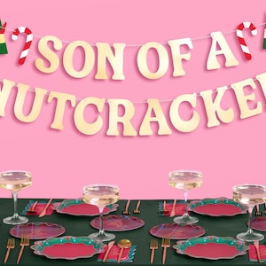 May include: A festive table setting with a pink background and a green tablecloth. Gold banner reads "SON OF A NUTCRACKER". The table is set with red and green plates, gold cutlery, and champagne glasses, creating a holiday theme.