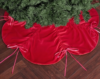 May include: A vibrant red Christmas tree skirt made of soft velvet. The skirt has a scalloped edge and is adorned with delicate pink ribbon ties. The skirt is placed around a green Christmas tree.