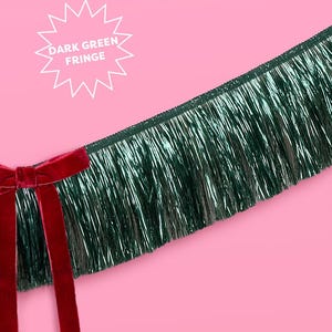 Puede incluir: Flecos verde oscuro con un lazo de terciopelo rojo sobre un fondo rosa. Los flecos tienen un aspecto brillante y met&aacute;lico. El texto en forma de estrella blanca dice "DARK GREEN FRINGE".