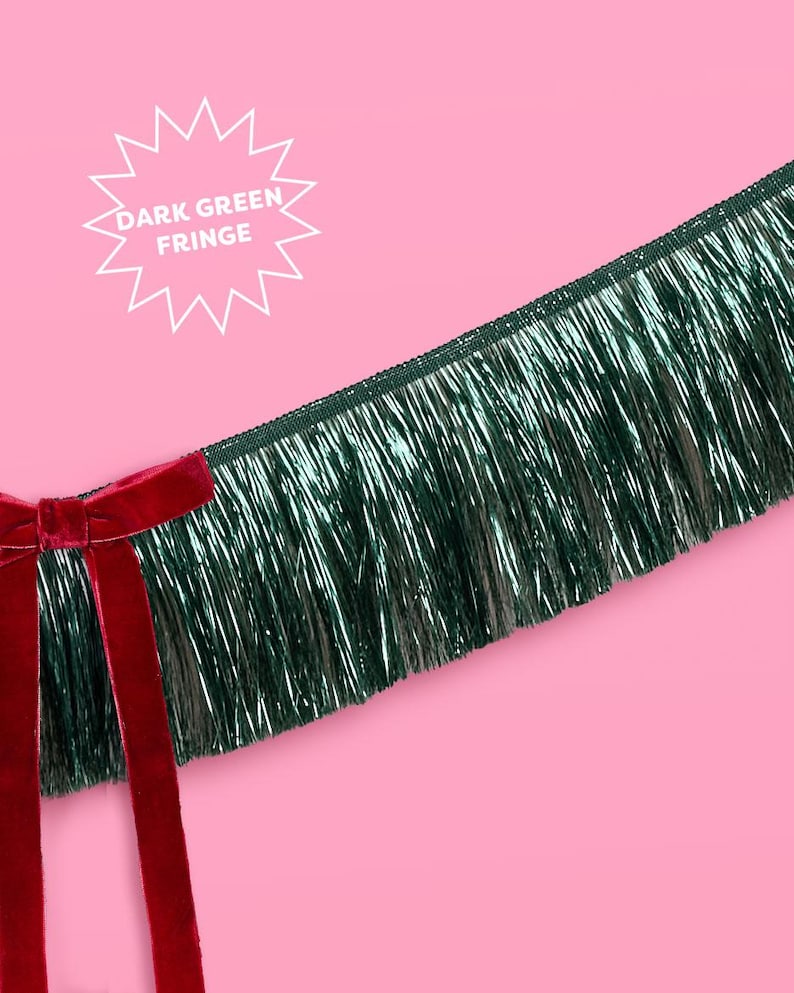 Puede incluir: Flecos verde oscuro con un lazo de terciopelo rojo sobre un fondo rosa. Los flecos tienen un aspecto brillante y met&aacute;lico. El texto en forma de estrella blanca dice "DARK GREEN FRINGE".