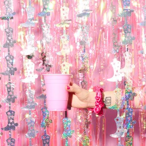 May include: A pink plastic cup in front of a pink and silver iridescent background with silver boot cutouts.