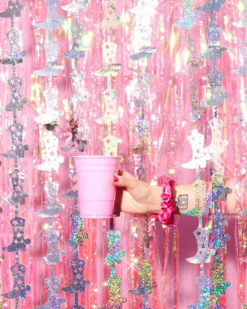 May include: A pink plastic cup in front of a pink and silver iridescent background with silver boot cutouts.