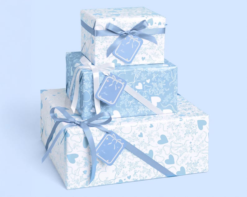May include: Three gift boxes stacked, wrapped in white paper with a blue heart and snowflake pattern. Each box is tied with a blue ribbon and bow, and has a matching tag. The background is a light blue.