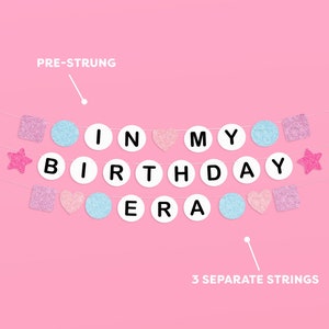 May include: A pink and blue pre-strung banner with the words "In My Birthday Era" in white letters on white circles. The banner includes pink and blue glitter stars, hearts, and squares.
