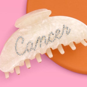 May include: White plastic hair claw with a rhinestone design that spells out "Cancer".