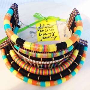 May include: Multi-layered colorful fabric necklace with black, yellow, orange, turquoise, pink, and gold accents. The necklace is made of multiple strands of fabric and has a chunky, bold design. The necklace is accompanied by a yellow tag with the text "Life is too short to wear boring jewelry".