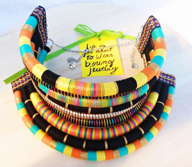 May include: Multi-layered colorful fabric necklace with black, yellow, orange, turquoise, pink, and gold accents. The necklace is made of multiple strands of fabric and has a chunky, bold design. The necklace is accompanied by a yellow tag with the text "Life is too short to wear boring jewelry".