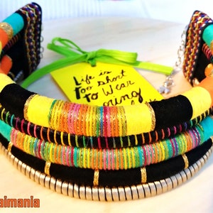 May include: A colorful multi-layered necklace with black fabric, yellow, orange, turquoise, and pink threads, and gold accents. The necklace is decorated with silver beads and has a quote card that reads "Life is short to wear boring things".