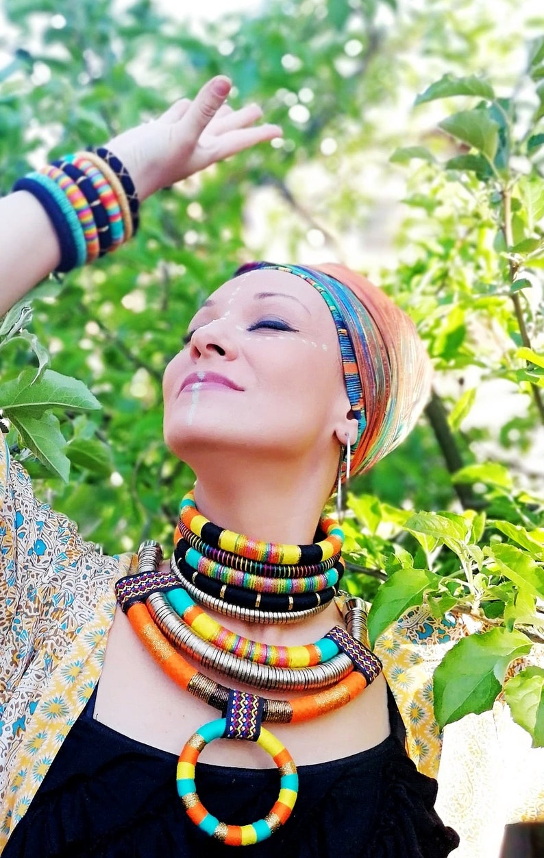 May include: A woman wearing a colorful head wrap and a layered necklace with a large, circular pendant. The necklace is made of various materials, including fabric, metal, and beads. The woman is wearing a black top and a floral patterned shirt.