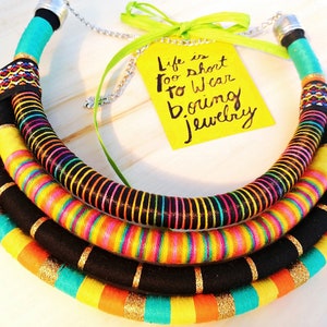 May include: A colorful multi-layered necklace made of fabric and thread. The necklace is black, yellow, orange, turquoise, and pink. The necklace is in the shape of a circle and has a quote on a yellow tag that reads "Life is too short to wear boring jewelry".