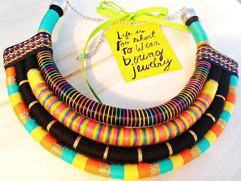 May include: A colorful multi-layered necklace made of fabric and thread. The necklace is black, yellow, orange, turquoise, and pink. The necklace is in the shape of a circle and has a quote on a yellow tag that reads "Life is too short to wear boring jewelry".