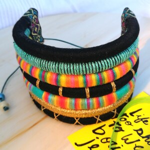 May include: A multi-layered fabric necklace with black, green, yellow, orange, pink, and blue stripes. The necklace is decorated with gold thread and has a black fabric backing.
