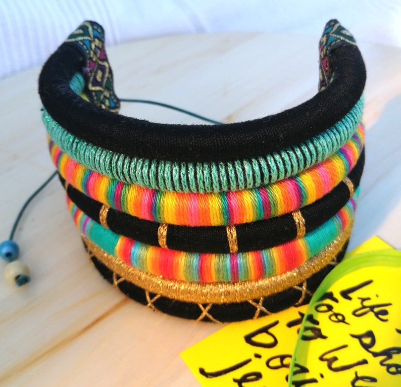 May include: A multi-layered fabric necklace with black, green, yellow, orange, pink, and blue stripes. The necklace is decorated with gold thread and has a black fabric backing.
