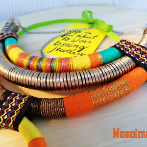 May include: A colorful necklace with multiple strands of fabric and metal beads. The fabric strands are orange, yellow, turquoise, and black with geometric patterns. The metal beads are silver and arranged in a circular pattern. The necklace is on a wooden surface with a yellow tag that says "Why is it so hard to wear boring jewelry?"