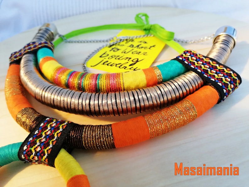 May include: A colorful necklace with multiple strands of fabric and metal beads. The fabric strands are orange, yellow, turquoise, and black with geometric patterns. The metal beads are silver and arranged in a circular pattern. The necklace is on a wooden surface with a yellow tag that says "Why is it so hard to wear boring jewelry?"