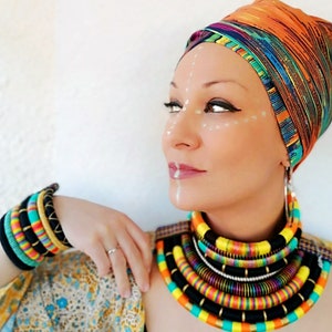 May include: A woman wearing a colorful head wrap and a multi-layered necklace made of colorful cords. She is also wearing several bracelets on her wrist.
