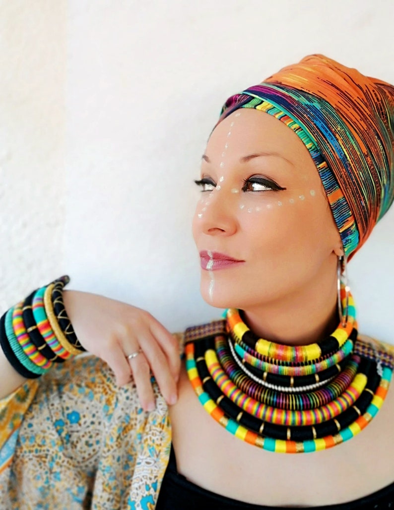 May include: A woman wearing a colorful head wrap and a multi-layered necklace made of colorful cords. She is also wearing several bracelets on her wrist.