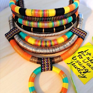 May include: Multi-layered colorful necklace with a bright orange, yellow, turquoise, and black color scheme. The necklace is made of woven fabric and metal. The necklace is accompanied by a smaller matching ring.