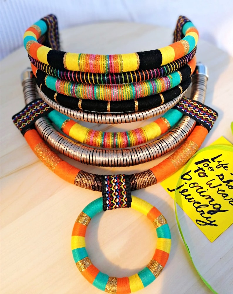 May include: Multi-layered colorful necklace with a bright orange, yellow, turquoise, and black color scheme. The necklace is made of woven fabric and metal. The necklace is accompanied by a smaller matching ring.