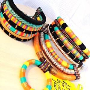 May include: A set of colorful, multi-layered necklaces made with woven fabric and metal. The necklaces are in shades of orange, yellow, turquoise, and black. The text on the tag reads "I love to fashion, too. What is your inclination?"