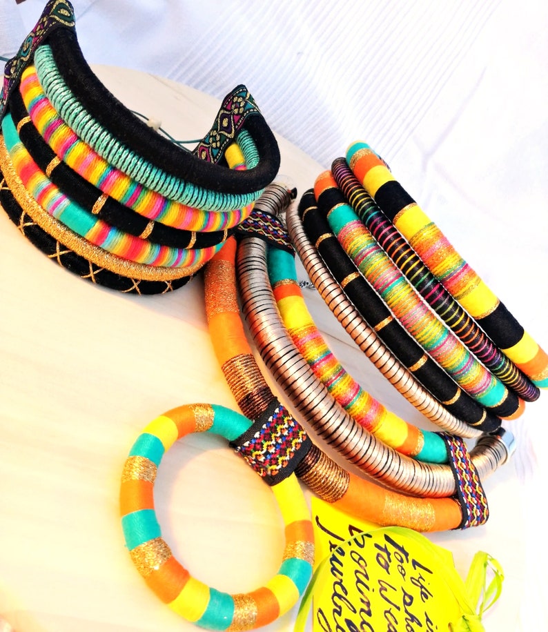 May include: A set of colorful, multi-layered necklaces made with woven fabric and metal. The necklaces are in shades of orange, yellow, turquoise, and black. The text on the tag reads "I love to fashion, too. What is your inclination?"