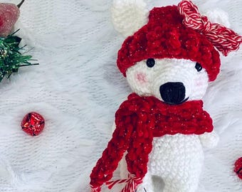 May include: A white crocheted bear toy wearing a red textured hat with a red and white striped bow, and a matching red textured scarf. The bear has black eyes, a black nose, and pink blush on its cheeks. The bear is made of yarn.