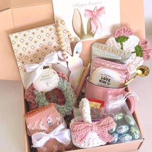 May include: An Easter gift box containing a variety of items. Features a card with bunny ears, a pink mug, a crochet basket with a pink bow, scrunchies, socks, and a bag of chocolates. The box is tied with a white ribbon.
