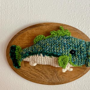 May include: A crochet fish wall hanging. The fish is blue, green, and white, mounted on an oval wooden plaque. The fish has a detailed design with fins and a tail. The background is a plain white wall.