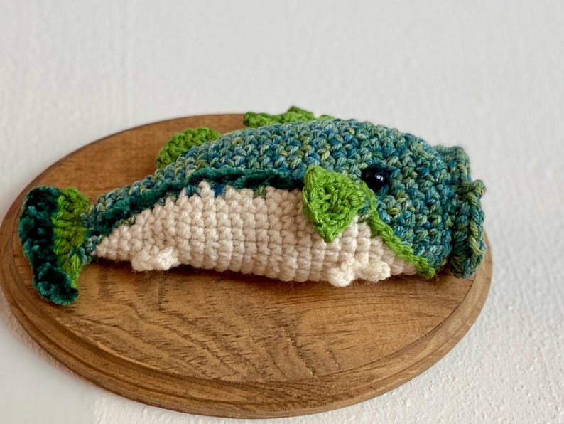 May include: A crocheted whale in blue, green, and white, displayed on a wooden plaque. The handmade whale has detailed stitching and a whimsical design, ideal for nautical-themed home decor.