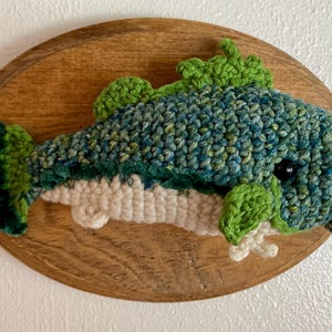 May include: A handmade crocheted fish, in shades of blue and green, mounted on an oval wooden plaque. The fish has detailed scales and fins, with a dark eye. A unique, handcrafted piece for the home.